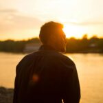A man stands by the river as the sun sets, creating a serene silhouette.